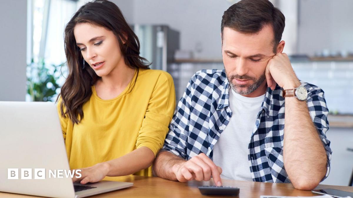 Couple sit at a kitchen table with the woman in a yellow jumper looking at a laptop and the man in a blue and white shirt