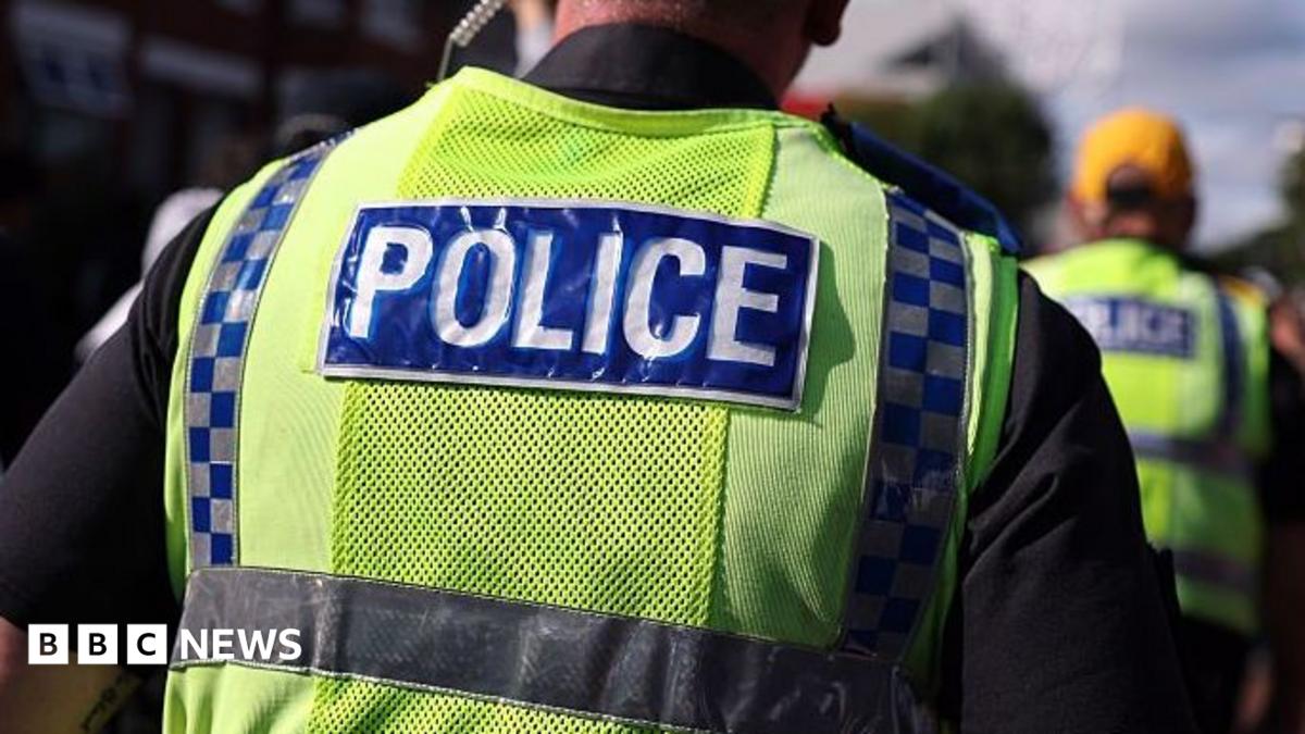 A police officer walks in a crowd alongside other officers. He has an ear piece and is wearing a fluorescent yellow tabard