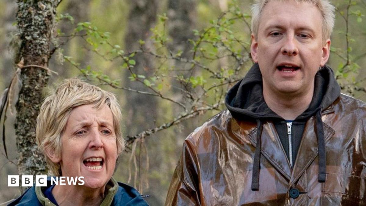 Julie Hesmondhalgh and Joe Lycett standing outdoors among trees and woodland vegetation, with one person wearing a blue