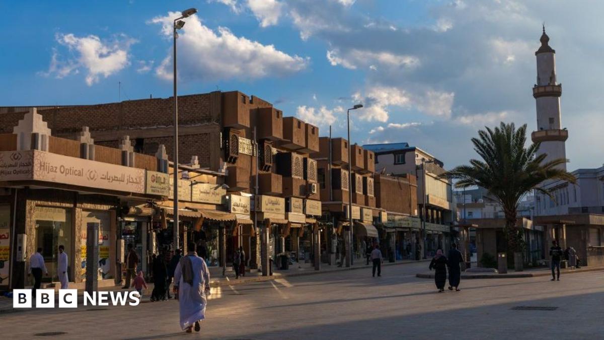 A broad street lined with low-rise shops stretches through a commercial district under late-afternoon light. Beige and brown