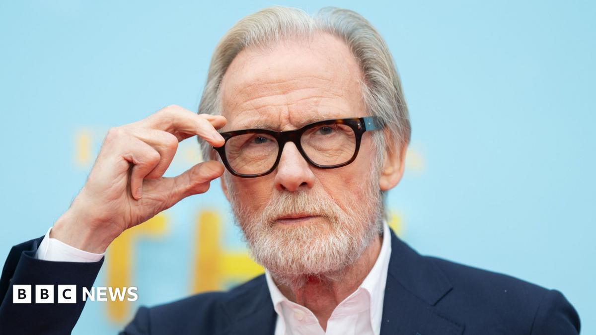 Bill Nighy in a dark suit with a white open necked shirt, with one hand on his black glasses
