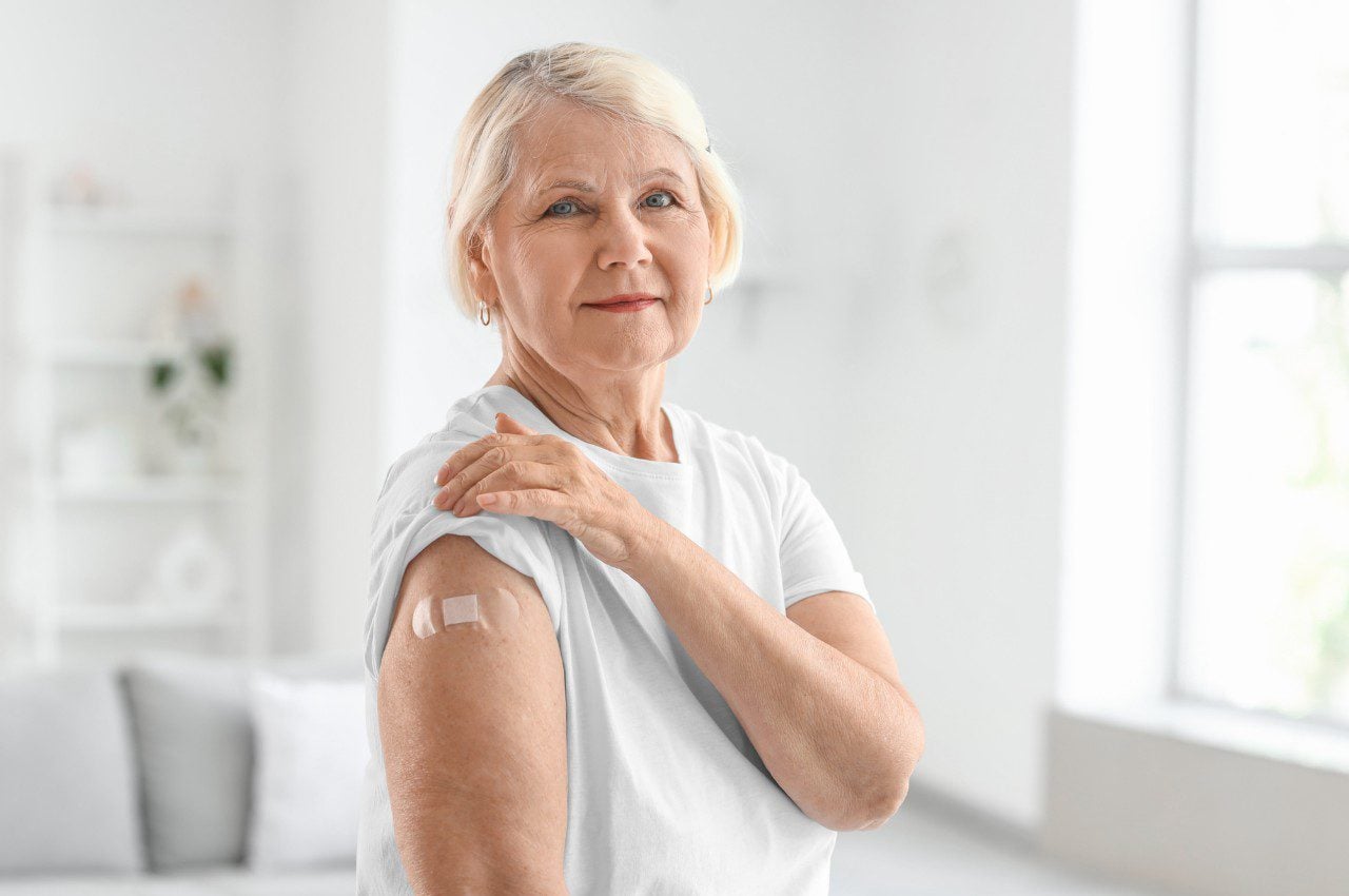 Older woman in a white shirt shows a small bandage on her upper arm after receiving a vaccination
