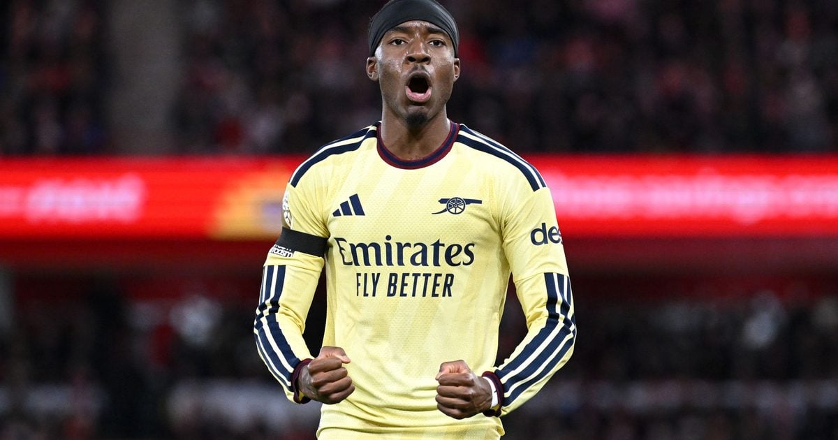 Arsenal winger Noni Madueke, in yellow and blue football kit, shouts and clenches both fists on the pitch in the Premier