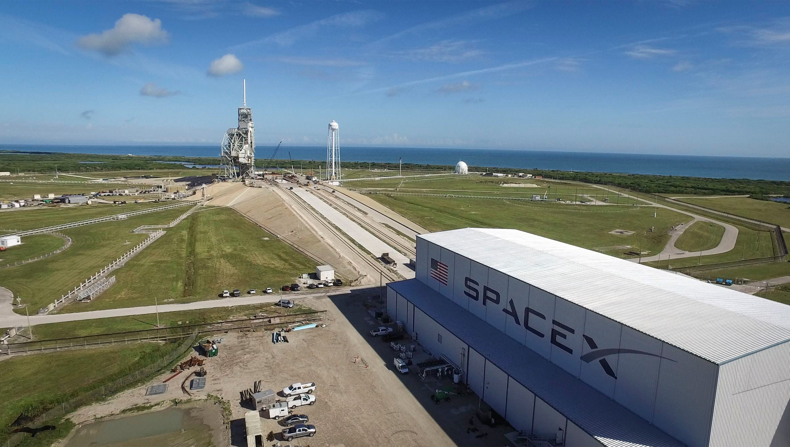 photo showing a seaside launch pad in the distance, with a boxy white building that says spacex on its side in the foreground