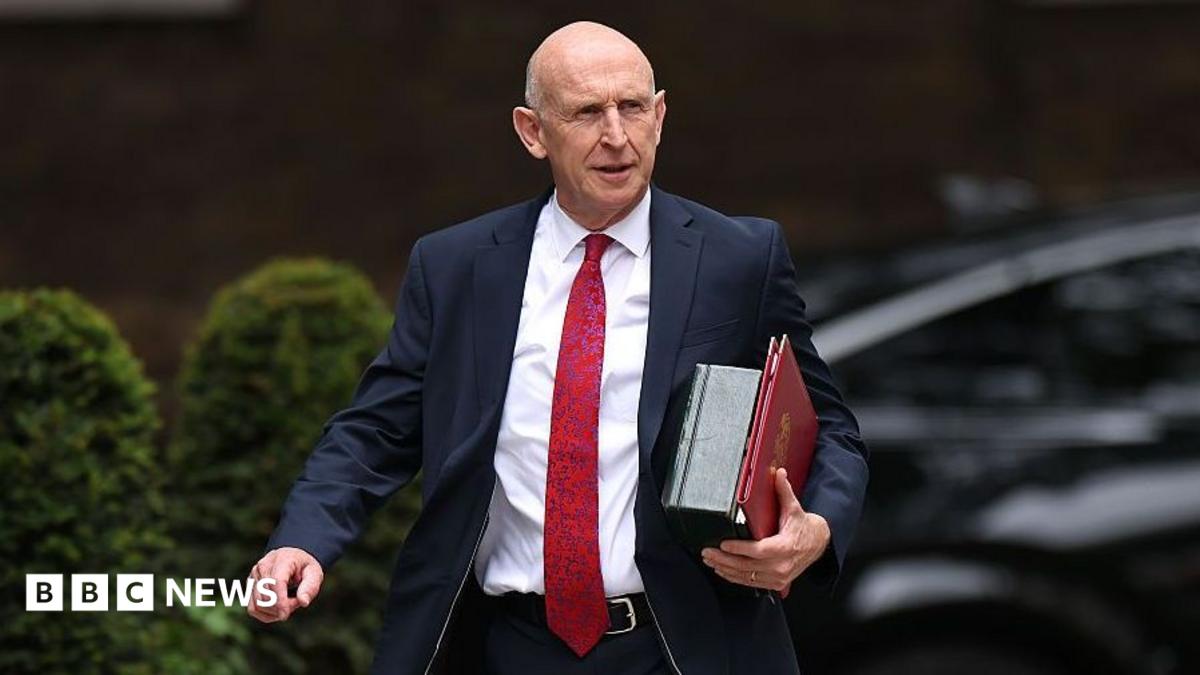 John Healey walks along Downing Street with files under his arm wearing a dark suit and a long red tie