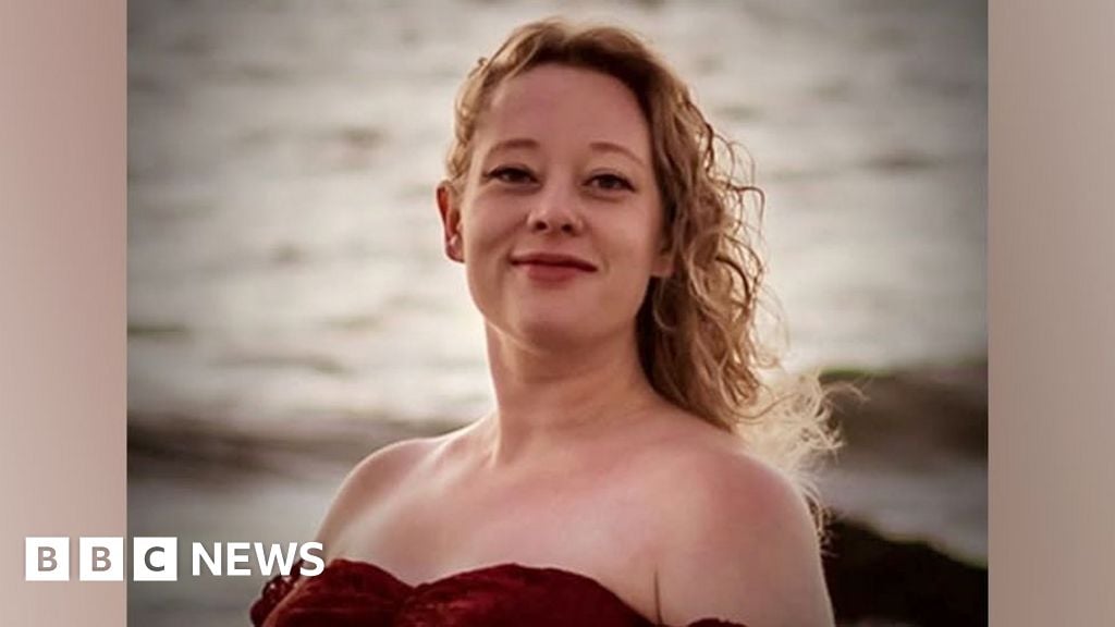 Renee Good has long, blonde, curly hair and is wearing a red off-the-shoulder top. The ocean is in the background