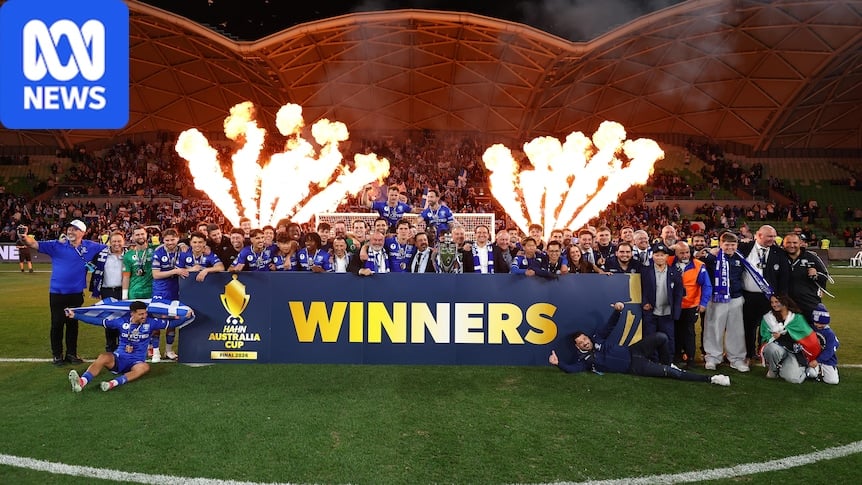 A group of South Melbourne soccer players and coaches gather together behind a sign saying 'Winners' as flames leap behind