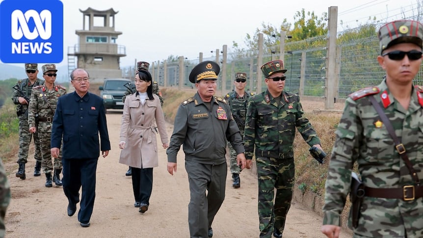A woman walks alongside a barbed wire fence with several men in army uniform
