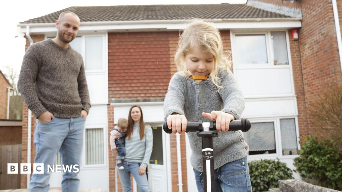 Family outside a house including a man, woman with a small child on her hip and another small child riding a scooter in the