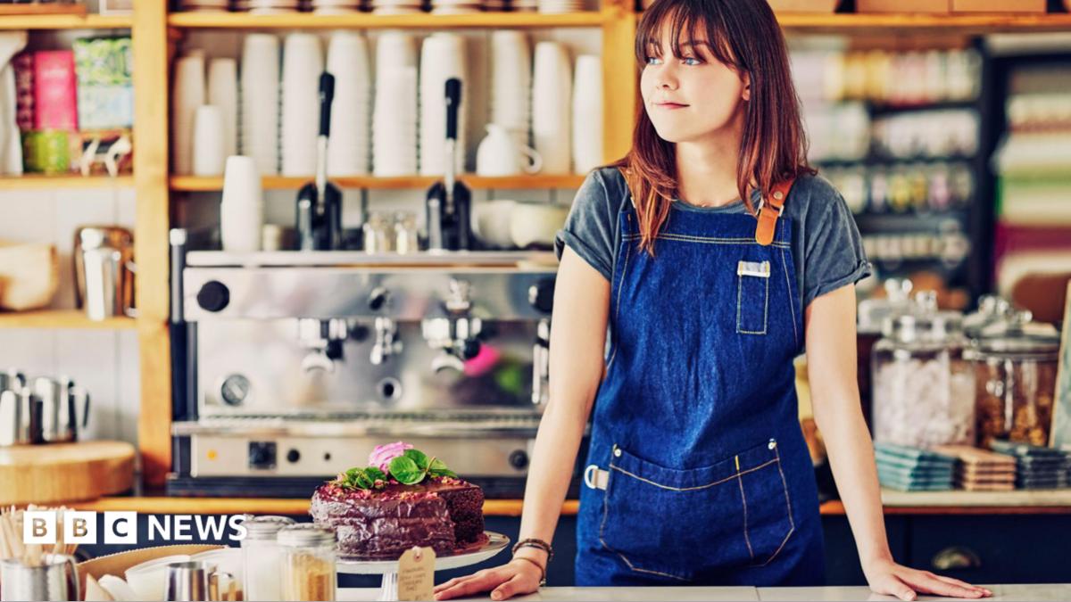 A young woman with long reddish-brown hair and wearing a dark blue apron stands behind the counter in a brightly-lit cafe