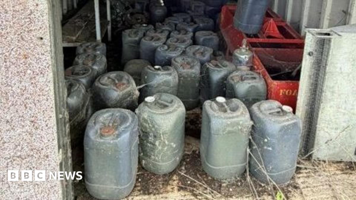A number of dark green containers inside what appears to be an open building. There are two red crates on one side. The