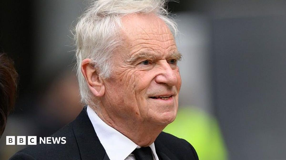 Jeffrey Archer is wearing a black suit and walking past the camera