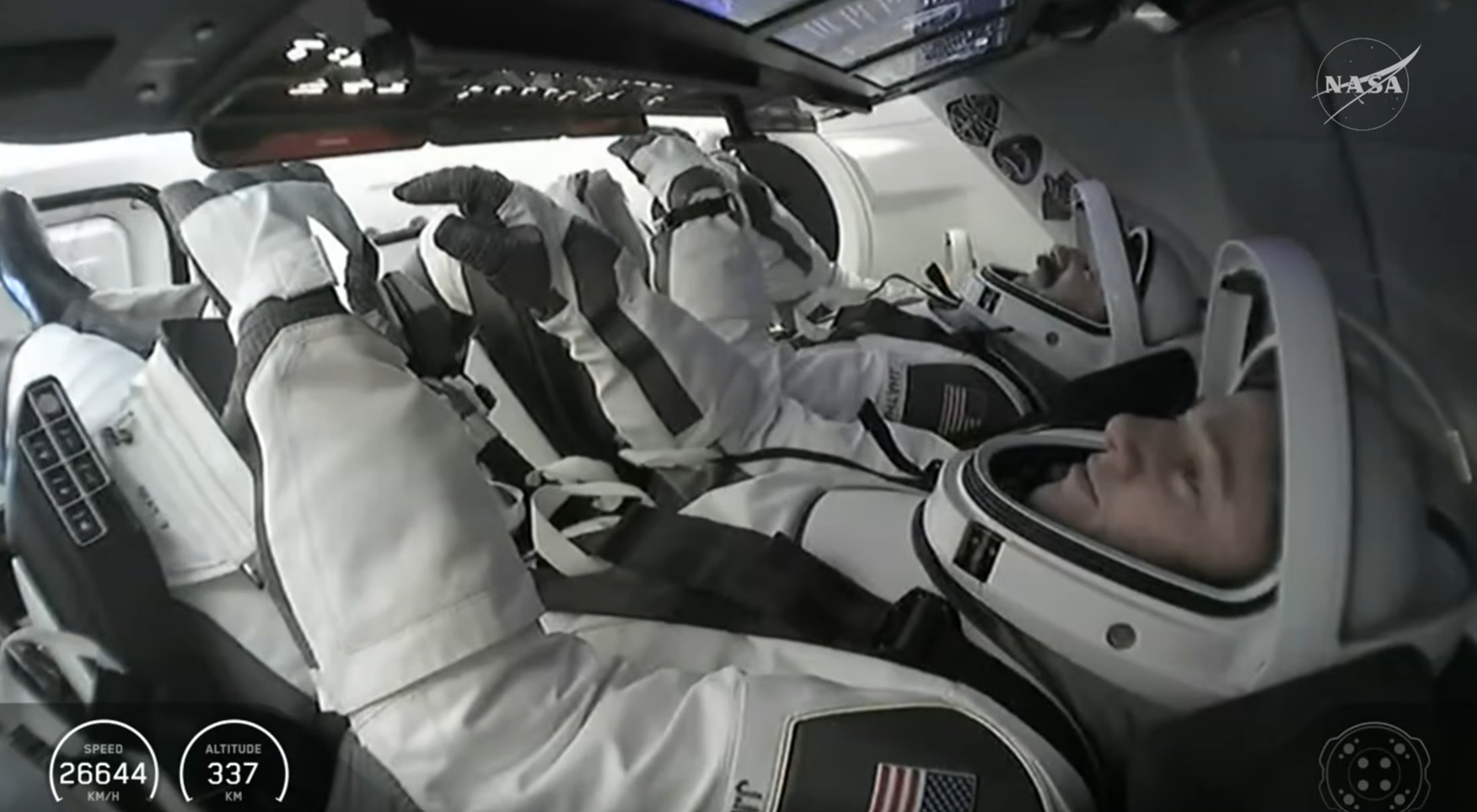 Image is inside a spacecraft with astronauts wearing white and black spacesuits