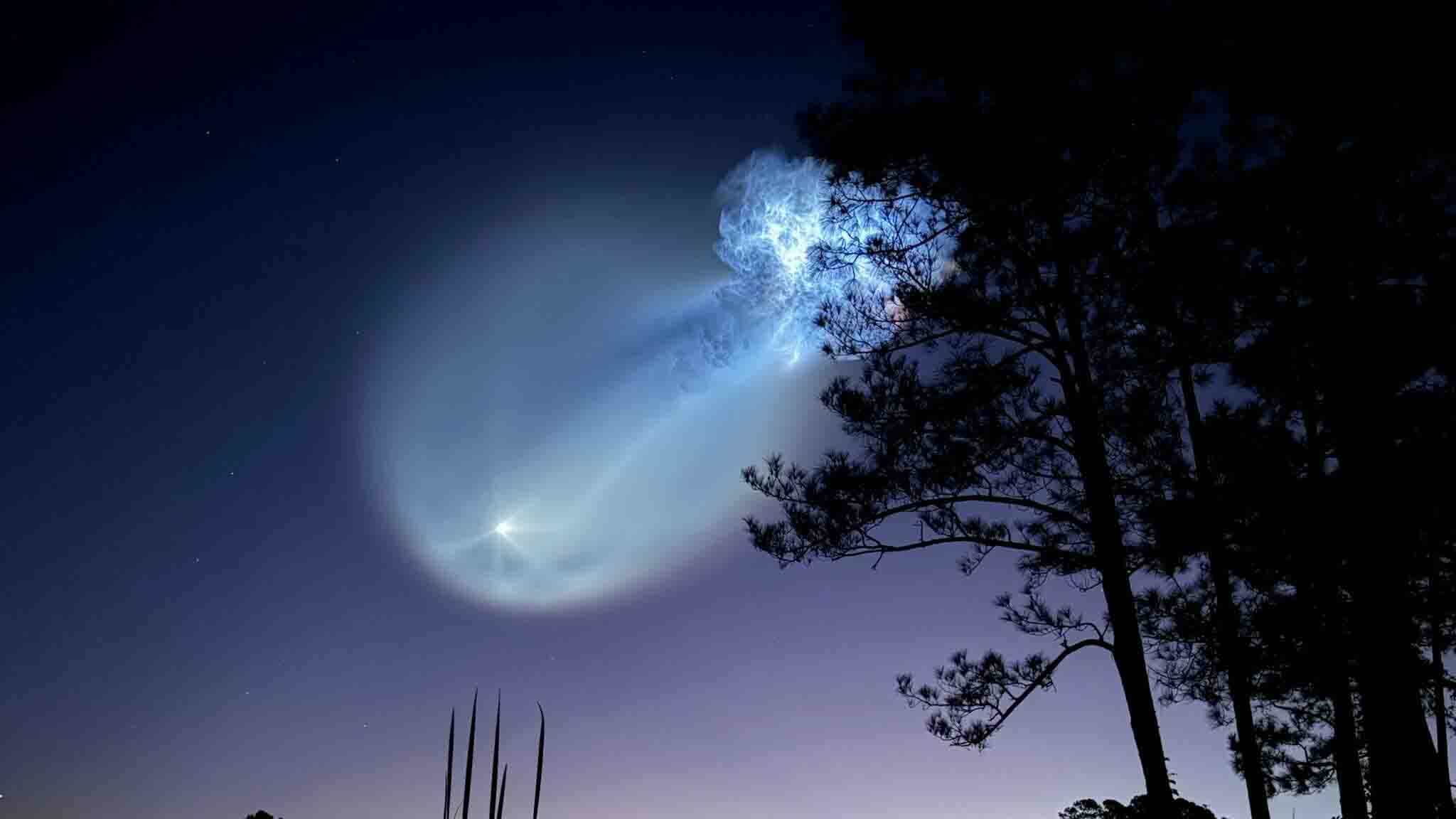 Glowing rocket exhaust trail forms a luminous spiral in a twilight sky above silhouetted pine trees